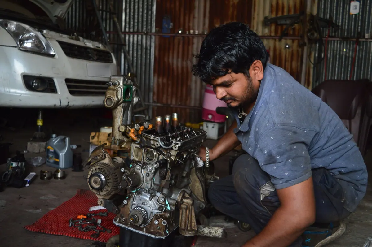 A mechanic crouched beside an engine block on the floor of a tin-roofed garage