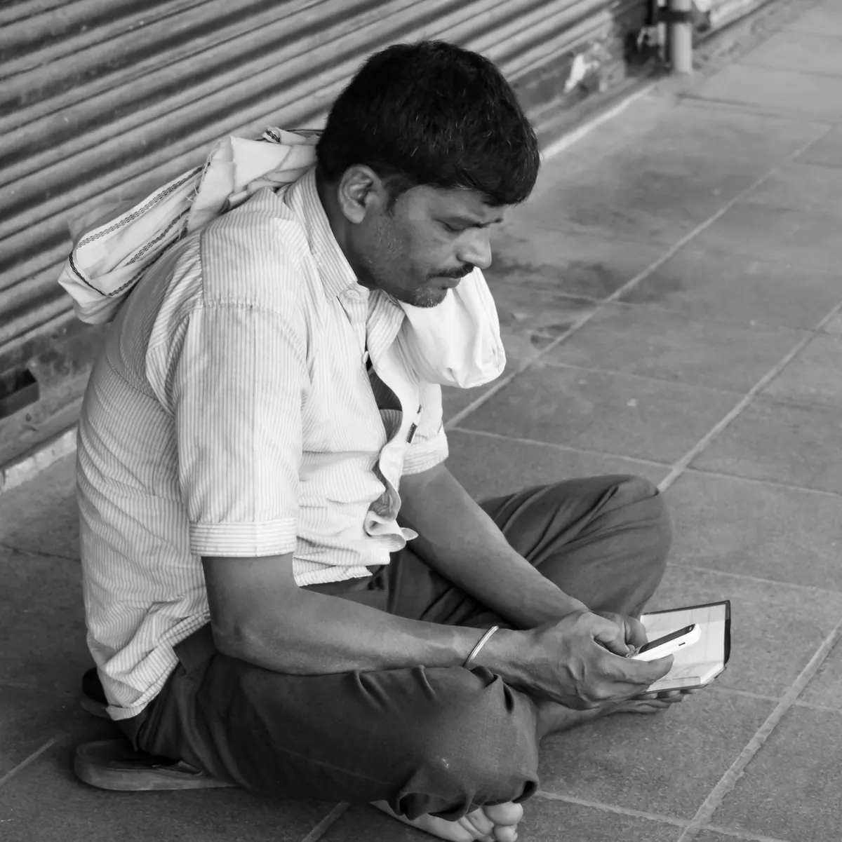 A man sitting cross-legged on a shop pavement, typing on a phone over a notebook