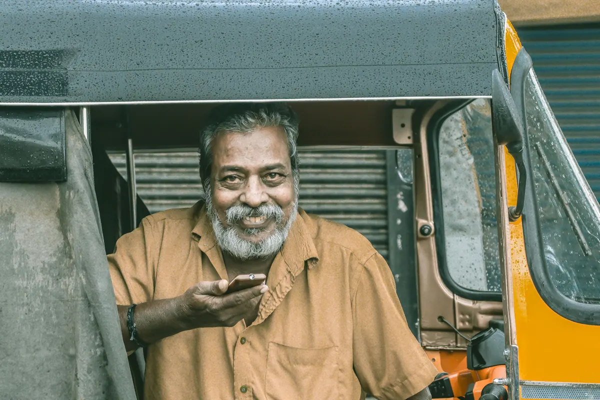 A man smiling with a phone in his hand, seated in an auto-rickshaw